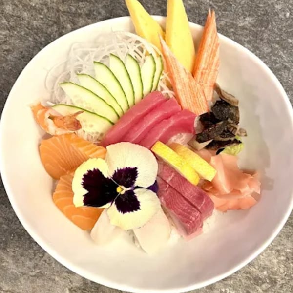 A colorful chirashi bowl with various sashimi, crab sticks, cucumber, and a pansy garnish.