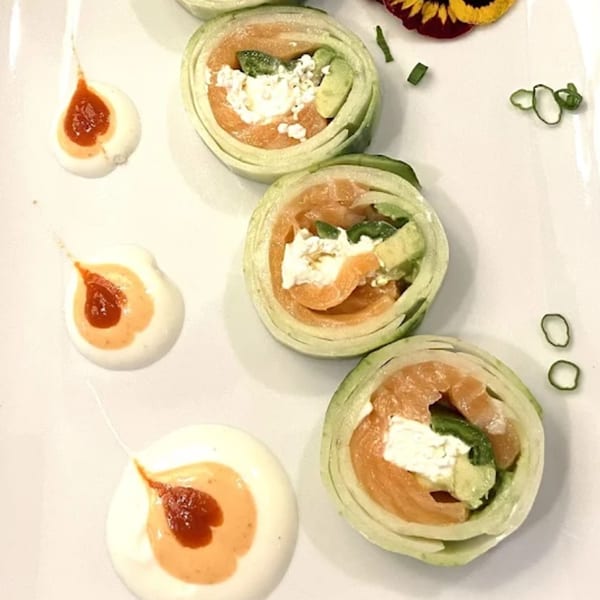 Three cucumber-wrapped sushi rolls with salmon, cream cheese, and avocado, garnished with sauces and herbs.