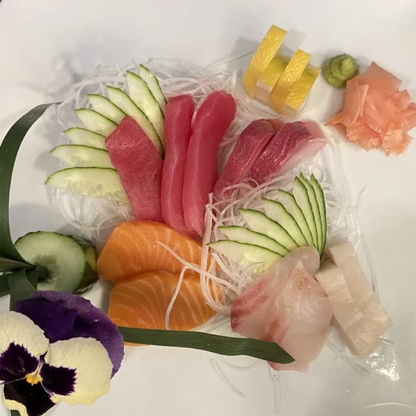 A colorful sashimi platter with tuna, salmon, and white fish, garnished with cucumber, lemon, and a pansy.