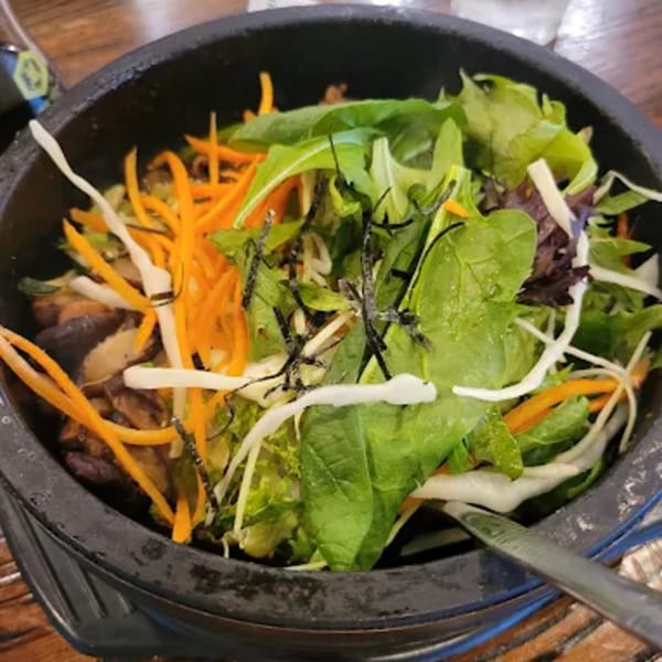A close-up of a colorful Korean bibimbap dish with mixed greens, carrots, and mushrooms in a black bowl.
